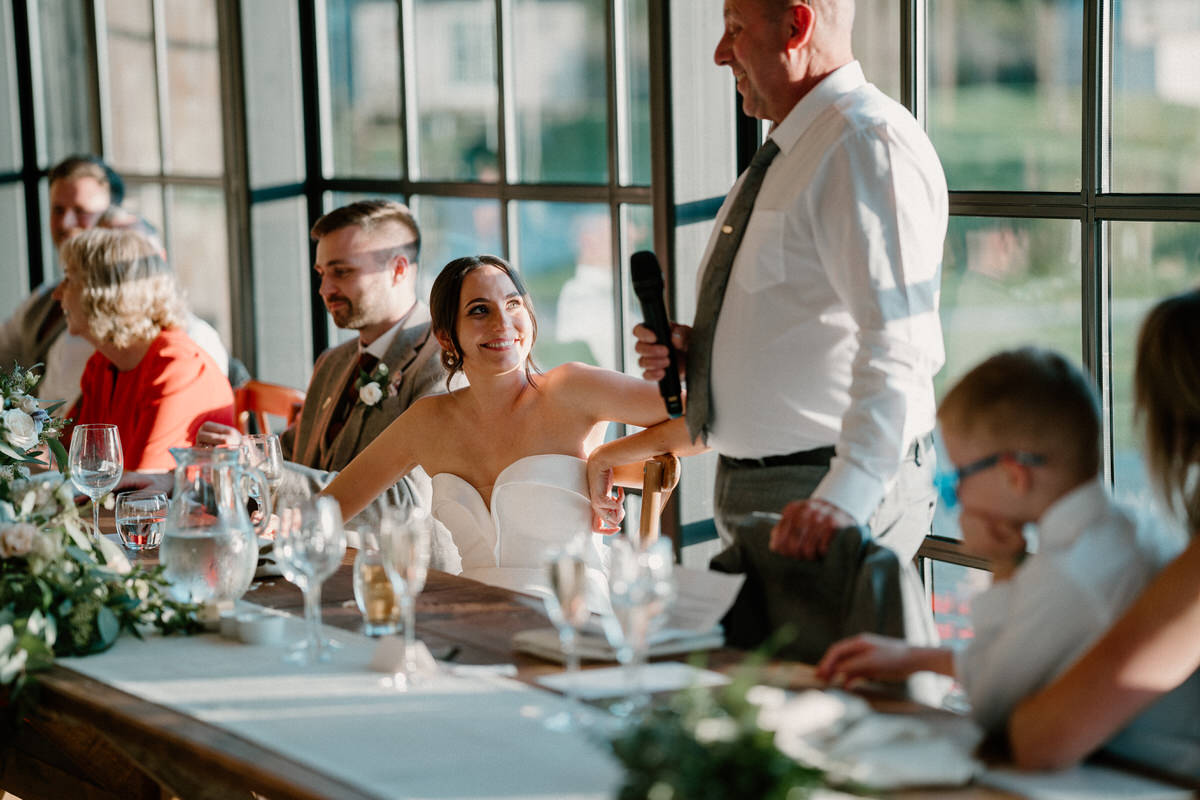 a bride looks on smiling as her father gives a wedding speech