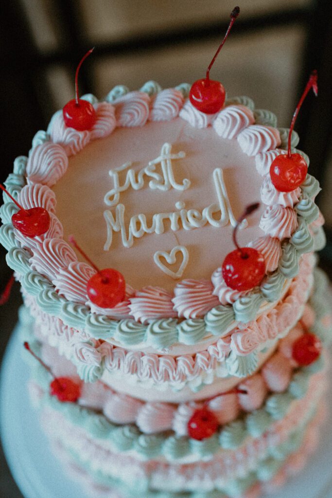 a retro white, pink and blue wedding cake, topped with cherries and 'just married' writing