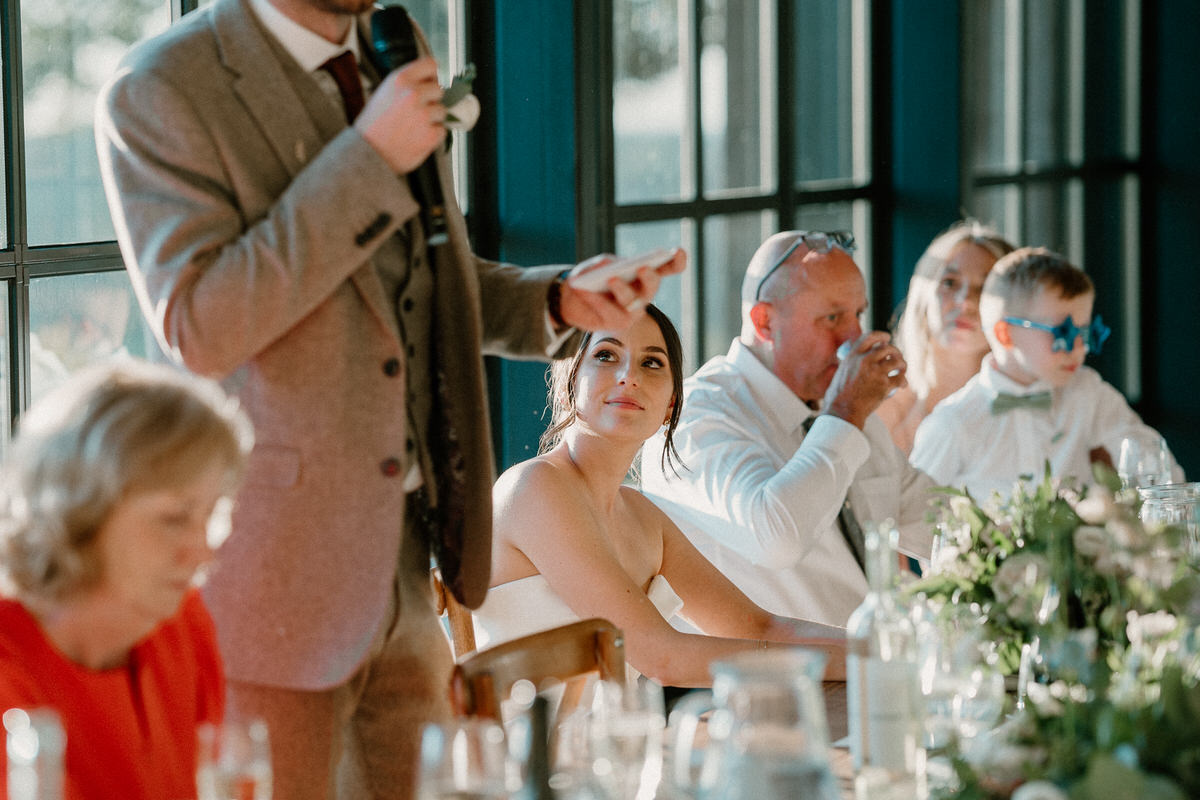 a bride looks on as her new husband gives a speech at their wedding breakfast