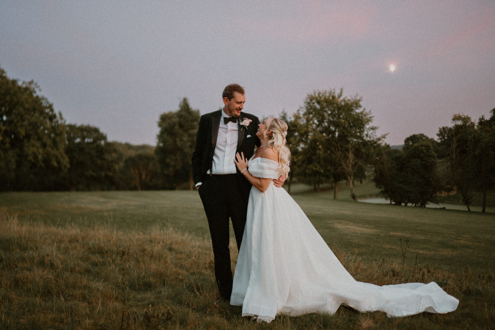 a bride hugging on to her new husbands arm, looking up at him with a pink sunset sky